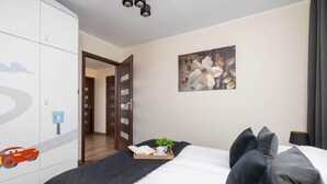 A hotel room with a bed, pillows, a bedside table with a book and a cup, a painting on the wall, and a wardrobe with a car design. - Spacious Apartment for 6 Persons | Balcony | 3 Bedrooms (Kraków)
