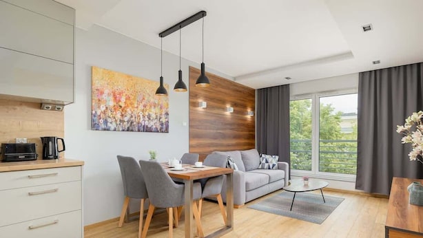 Un salon moderne comprenant un coin repas, un canapé et une table basse. On y trouve une grande fenêtre avec des rideaux, un tableau au mur, ainsi qu’un coin cuisine avec des placards et une machine à café.