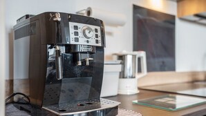 Una máquina de espresso DeLonghi sobre una encimera de cocina, con una cafetera y una vitrina de vidrio al fondo.