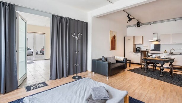 Habitación de hotel moderna con cama, zona de estar con sofá y mesa, kitchenette y vista al estacionamiento.