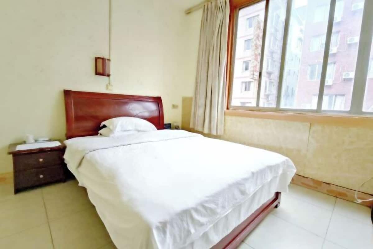 A bedroom with a bed, a nightstand, a lamp, a window with curtains, and a view of buildings outside.
