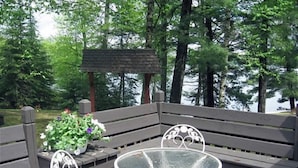 A wooden deck with a glass table and two chairs, surrounded by trees and a body of water. - Stepaway Vacation Rentals- Crazy Loon Lodge (St.Germain)