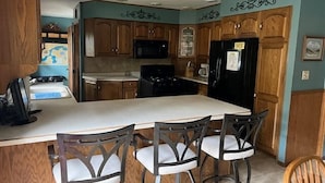 A kitchen with wooden cabinets, a black refrigerator, a stove, and a large island with bar stools. - Stepaway Vacation Rentals- Crazy Loon Lodge (St.Germain)