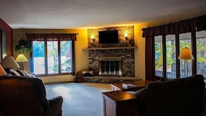 A living room with a fireplace, two windows with curtains, and a television mounted above the fireplace. - Stepaway Vacation Rentals- Crazy Loon Lodge (St.Germain)