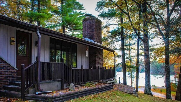 A rustic cabin with a brick chimney, wooden porch, and a view of a lake surrounded by trees. - Stepaway Vacation Rentals- Crazy Loon Lodge (St.Germain)