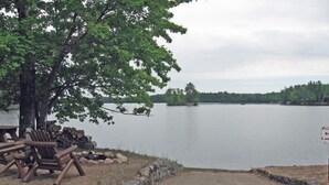 A lakeside picnic area with wooden chairs and a stack of firewood, a paved path leading to the water, and a sign indicating private beach access. - Stepaway Vacation Rentals- Cla-Rie Cabin (St.Germain)