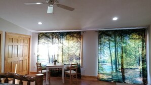 Una habitación con una mesa de comedor, sillas, un ventilador de techo y un amplio ventanal con vistas a un bosque.