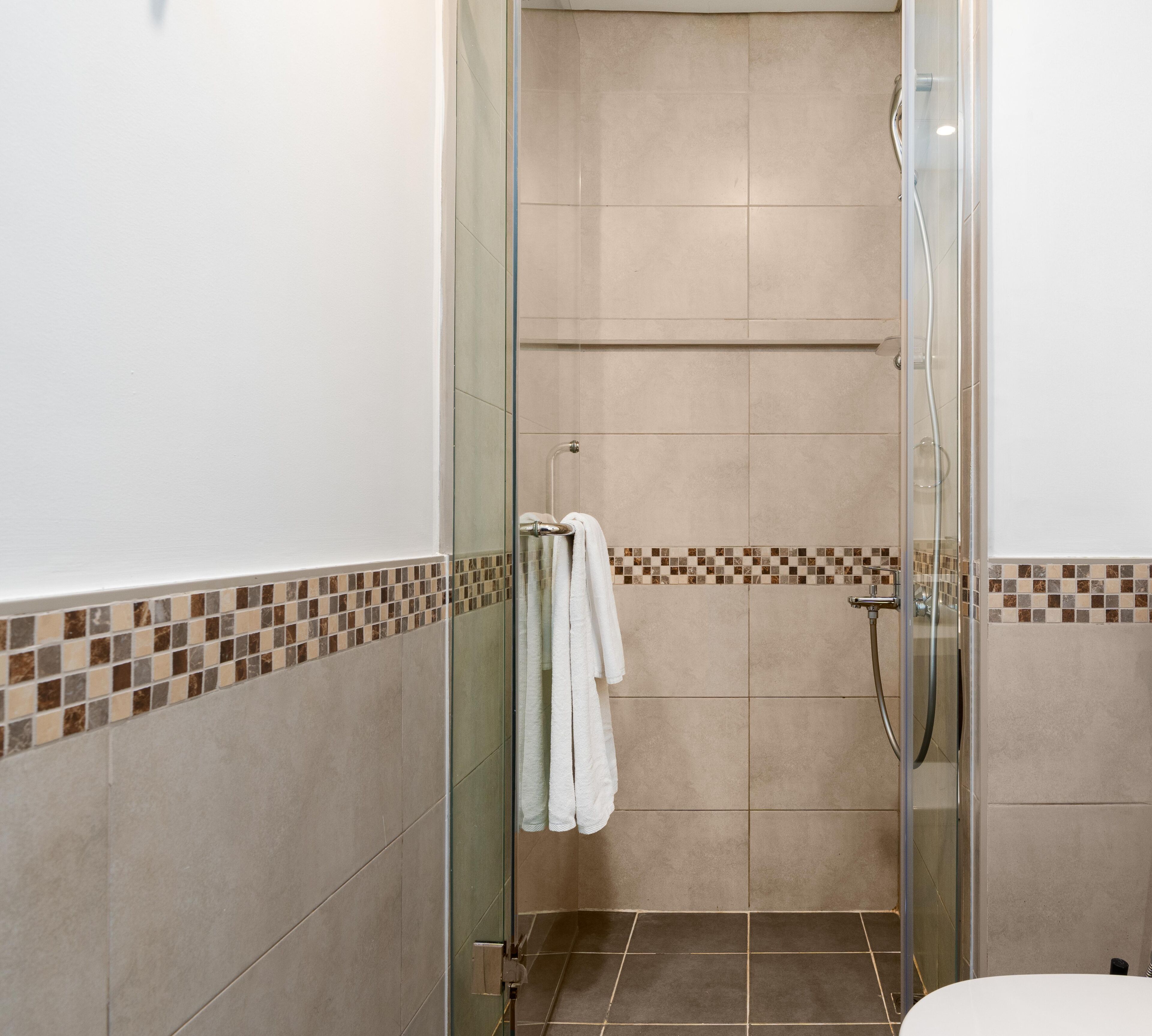 A glass-enclosed shower with a tiled floor and walls, a towel hanging on the door, and a small shelf at the bottom.