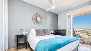 Un dormitorio moderno con cama, mesitas de noche, ventilador de techo, espejo montado en la pared y vistas a la ciudad a través de un ventanal.