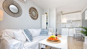 Un salón moderno con un sofá blanco, una mesa de comedor con un frutero, y una cocina al fondo.