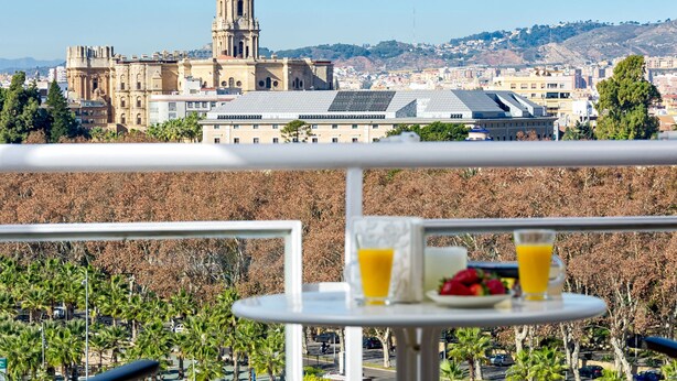 Un balcón con una mesa dispuesta para el desayuno, con vistas a un paisaje urbano que incluye un edificio destacado y colinas al fondo.