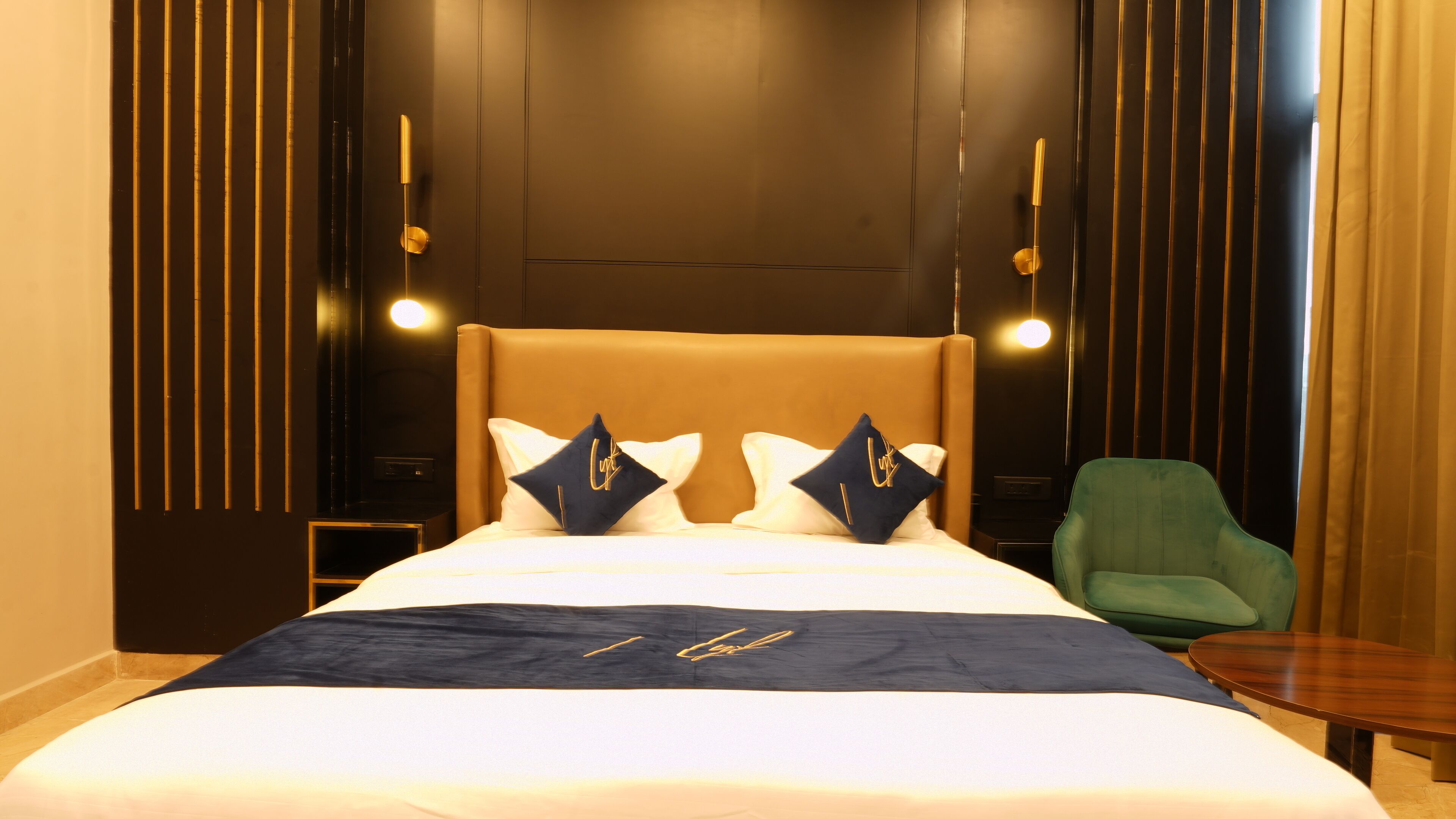Una habitación de hotel con una cama grande, dos mesitas de noche, un sillón verde y una pequeña mesa de madera.