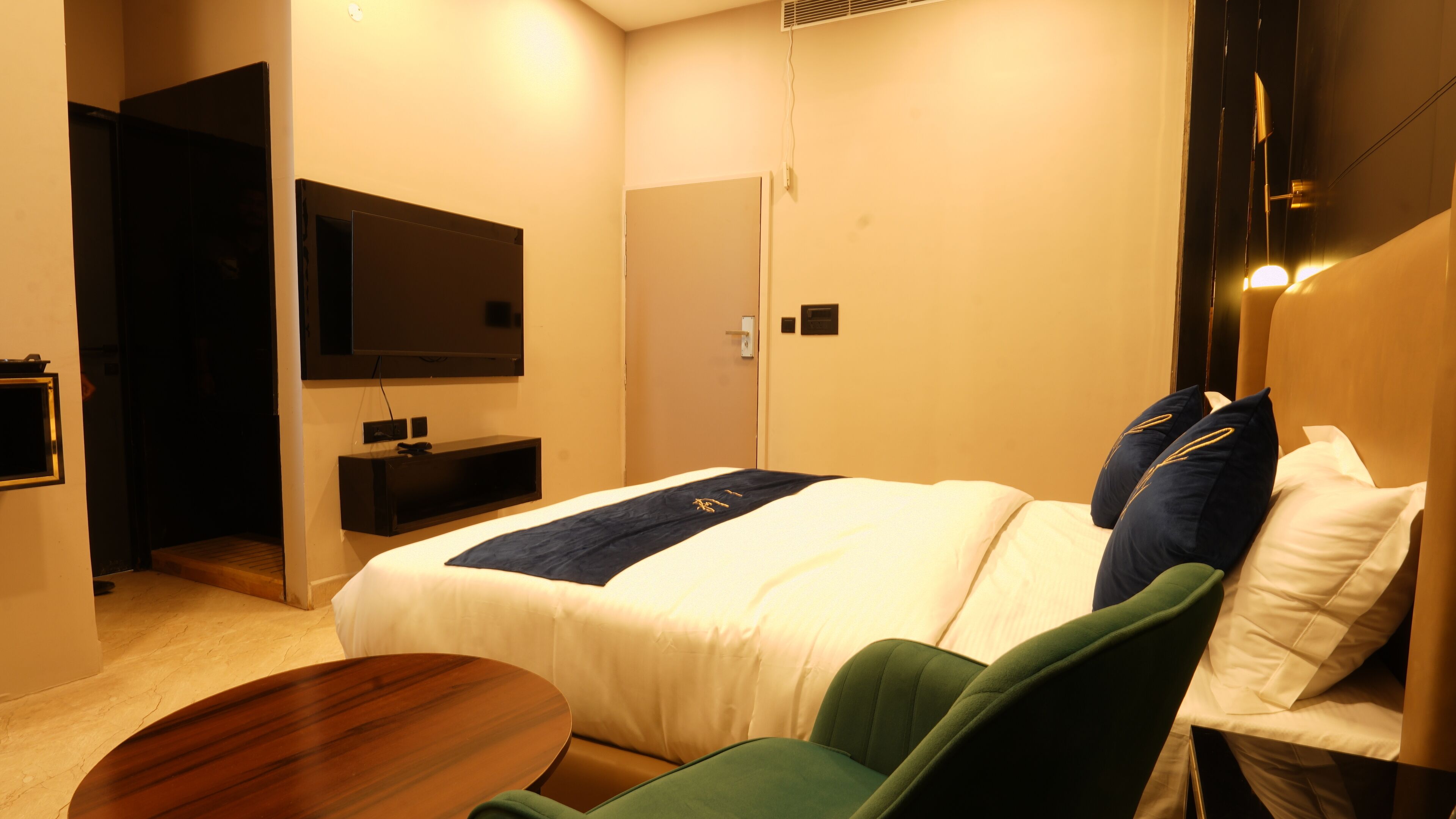 Habitación de hotel con cama, televisor montado en la pared, mesita de noche y un sillón verde.