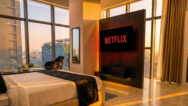 A modern hotel room with a large bed, a TV mounted on the wall, and a view of the cityscape through large windows. - Aquila Grand Suites (Noida)