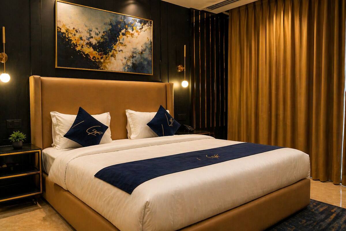 A hotel room with a large bed, a headboard, two bedside tables, a nightstand, a potted plant, a painting, and a curtain.