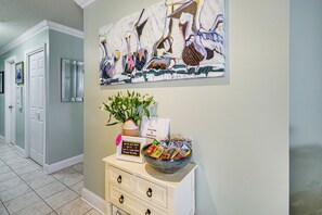 A hallway with a white door, a framed painting, a vase with flowers, a sign, and a bowl with bottled water. - Waterfront Biloxi Condo: Gulf-view Balcony! (Biloxi)
