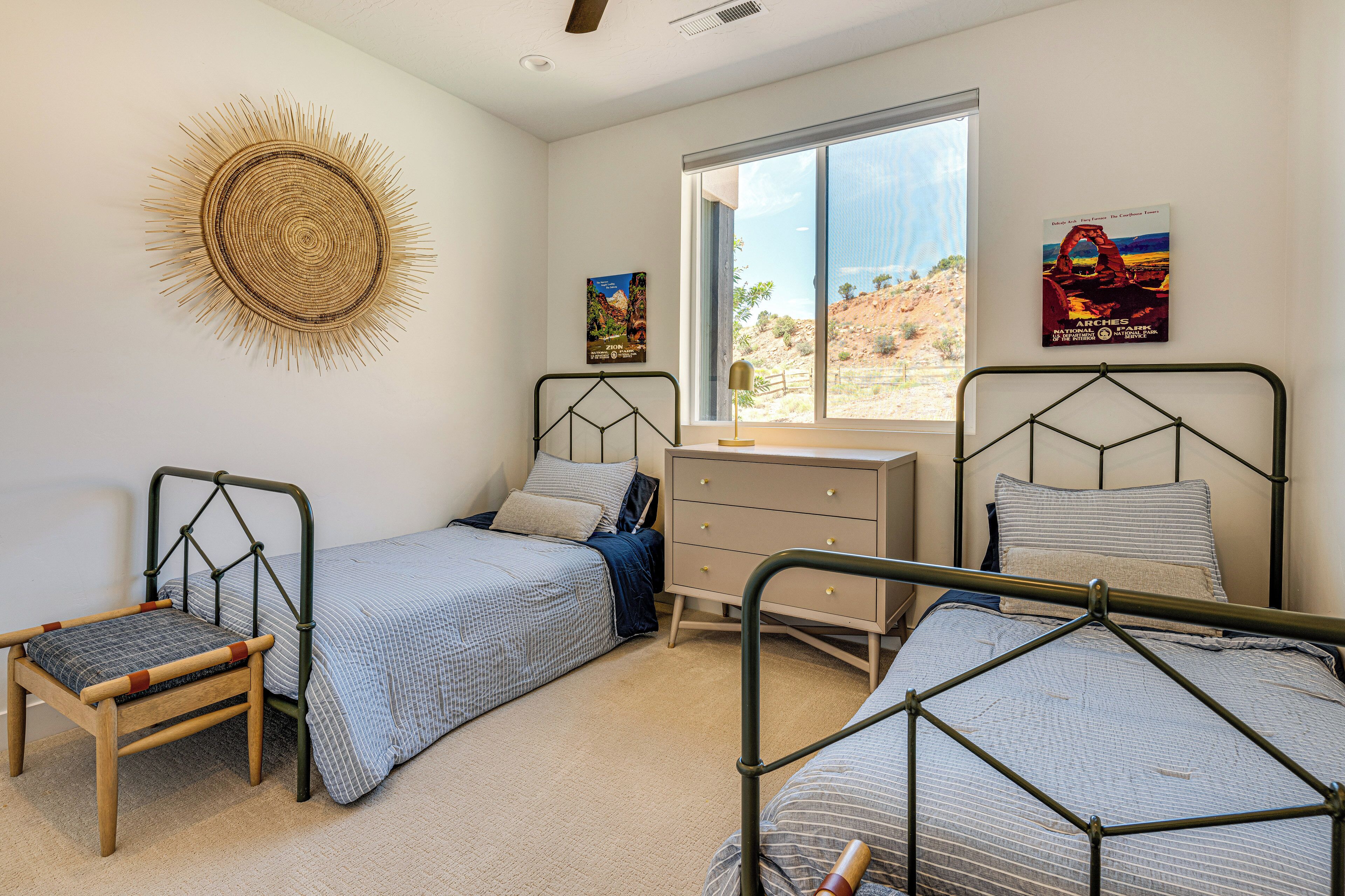A bedroom with two single beds, a wooden chair, a dresser, and a large window with a view of a desert landscape.