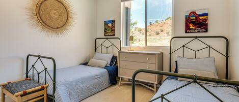 A bedroom with two single beds, a wooden chair, a dresser, and a large window with a view of a desert landscape.