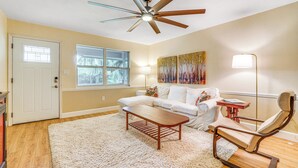 A living room with a ceiling fan, a white sofa, a wooden coffee table, a rocking chair, and a large window. - Half-mi to Riverwalk: Charming Sanford Retreat! (Sanford)