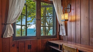 Una cabaña de madera con un banco y una mesa, una ventana con cortinas y una lámpara fijada a la pared.