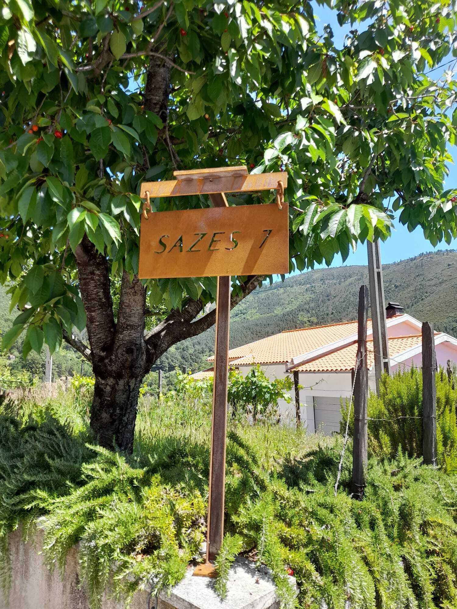 A rustic wooden sign with the text "SAZES 7" in a rural setting with a tree, greenery, and a building with a red roof in the background.