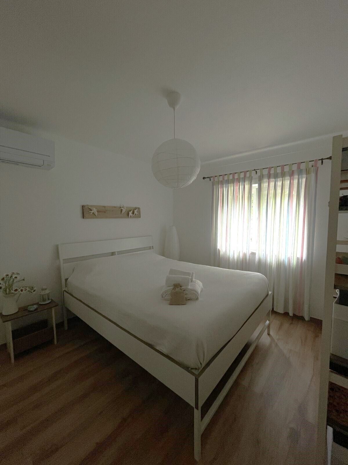 Ein Schlafzimmer mit einem Einzelbett, einem Nachttisch mit Lampe, einer Wand-Klimaanlage und einem Fenster mit Vorhängen.