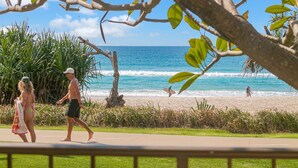 A beach with people walking and surfing, palm trees, and a clear view of the ocean.