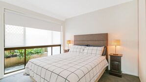 A bedroom with a large bed, two bedside tables with lamps, a window with a view of plants, and a balcony.