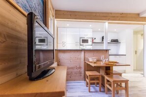 Moderni keittiö, jossa on valkoiset kaapit, mikroaaltouuni ja kahvinkeitin. Keittiössä on puinen ruokapöytä penkeillä sekä televisio puisen jalustan päällä.