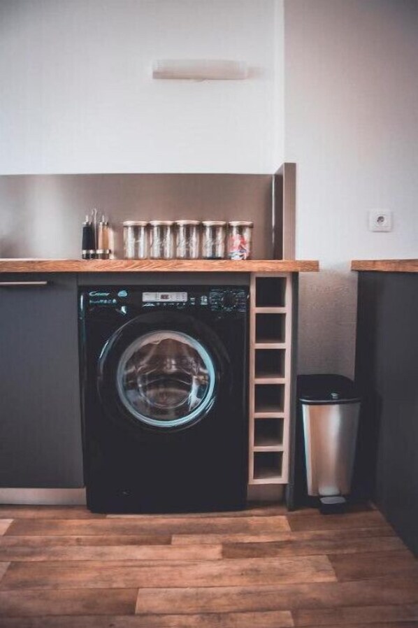 A compact laundry area with a front-loading washing machine, a small trash bin, and a wooden shelf with cans.