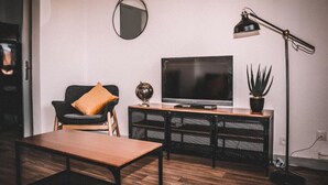 A modern living room with a flat-screen TV, a black chair with a brown cushion, a wooden coffee table, a black floor lamp, and a potted plant.