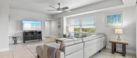 A spacious living room with a large sectional sofa, a ceiling fan, a flat-screen TV, and a window with blinds.