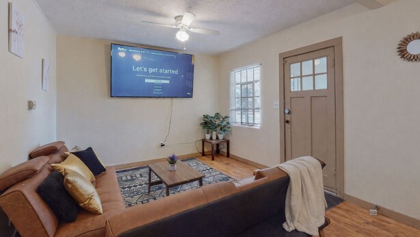 A living room with a sofa, a coffee table, a TV mounted on the wall, a ceiling fan, and a door.