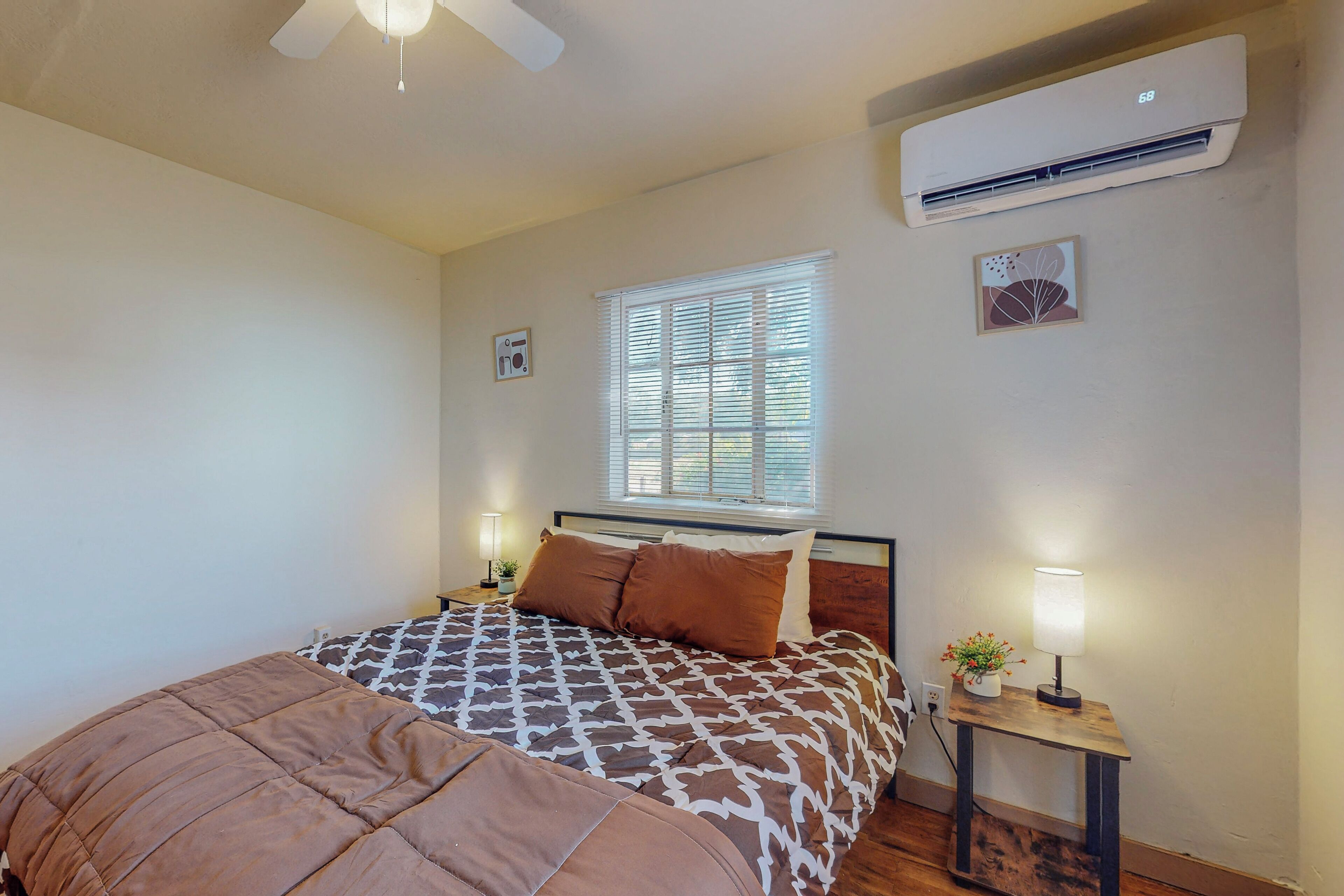 A bedroom with a bed, a bedside table with a lamp, a window with blinds, an air conditioner, and a ceiling fan.