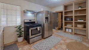 A compact kitchen with a gas stove, oven, and refrigerator, a potted plant, and a wooden shelving unit with baskets.