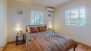 A bedroom with a bed, two bedside tables with lamps, an air conditioner, and two windows with blinds.