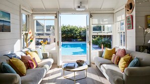 A sunlit living room with a view of a swimming pool, two sofas with colorful cushions, and a small table with a bowl. - Beachfront House w Pool Steam (Chichester)