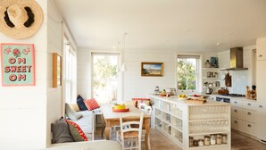 A bright, open-plan kitchen and dining area with white cabinetry, a wooden dining table, and a cozy seating area with cushions. - Beachfront House w Pool Steam (Chichester)