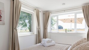 A bedroom with a bed, beige curtains, a window with an umbrella, and a view of trees. - Idyllic 3 Bed Country Cottage (Chichester)