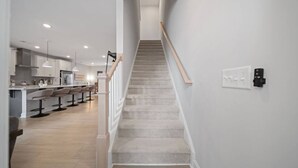 A modern kitchen with a central island, a dining area with bar stools, and a staircase with a wooden handrail. - Downtown Wake Forest Luxury Condo | Rooftop Terrace + EV Charger (Wake Forest)