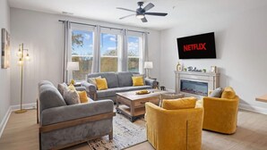 A modern living room with a grey sofa, yellow armchairs, a coffee table, and a fireplace. A ceiling fan and a TV mounted on the wall are also visible. - Downtown Wake Forest Luxury Condo | Rooftop Terrace + EV Charger (Wake Forest)