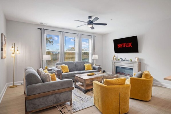 A modern living room with a grey sofa, yellow armchairs, a coffee table, and a fireplace. A ceiling fan and a TV mounted on the wall are also visible. - Downtown Wake Forest Luxury Condo | Rooftop Terrace + EV Charger (Wake Forest)