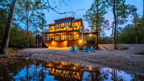 A modern two-story building with a glass facade, surrounded by trees and a gravel path leading to a seating area with chairs. - Island in the Stream Spacious Creekside Cabin - Wi-Fi, Hot Tub, Arcade (Broken Bow)