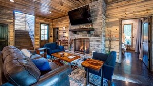 A log cabin interior with a fireplace, leather sofa, and a television mounted on the wall. - Island in the Stream Spacious Creekside Cabin - Wi-Fi, Hot Tub, Arcade (Broken Bow)