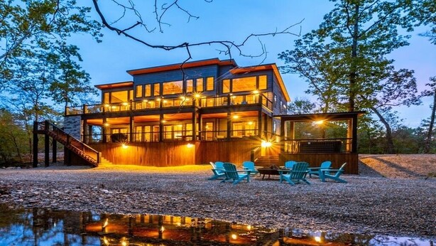 A modern two-story house with a deck and outdoor seating area, surrounded by trees and a body of water. - Island in the Stream Spacious Creekside Cabin - Wi-Fi, Hot Tub, Arcade (Broken Bow)