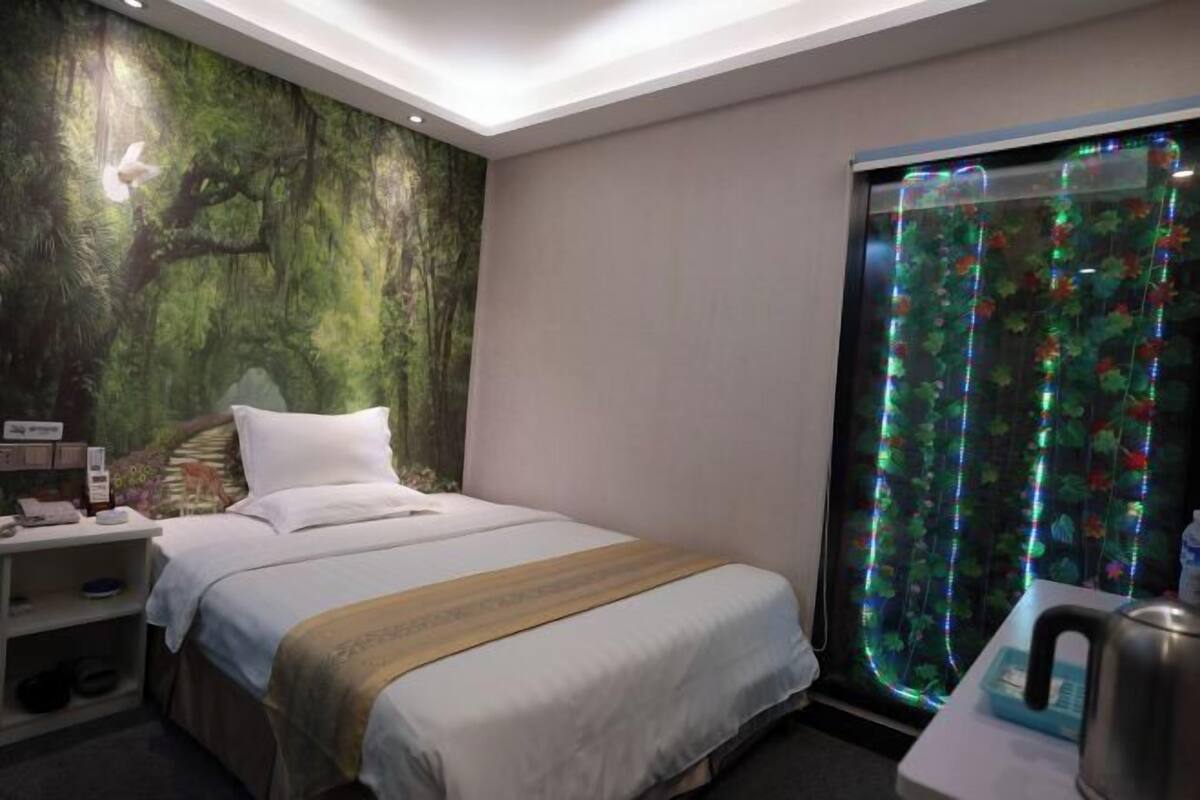 A hotel room with a bed, a nightstand, a desk with a kettle, and a window with a curtain.