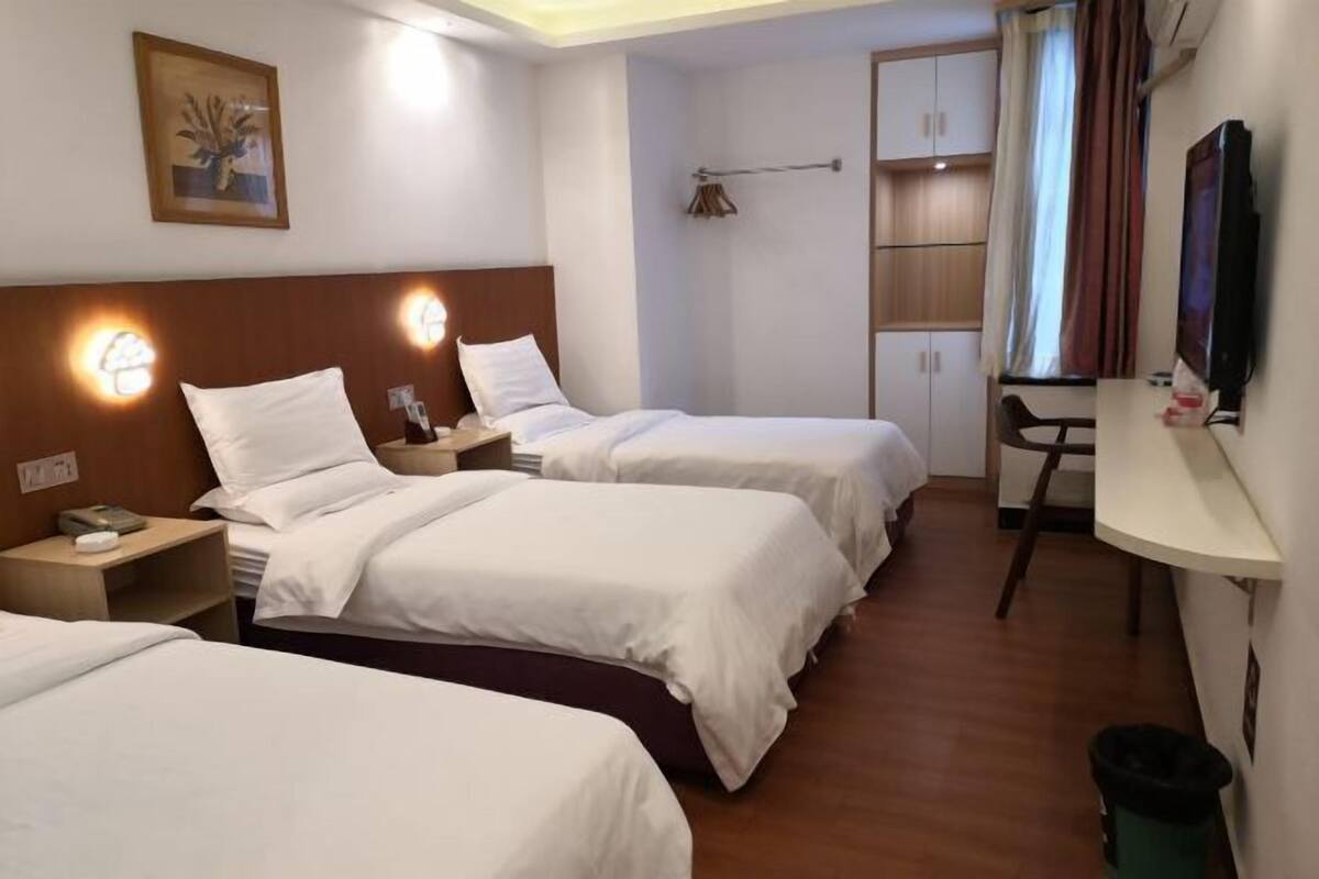 A hotel room with three single beds, a desk, a chair, a TV, and a trash bin.