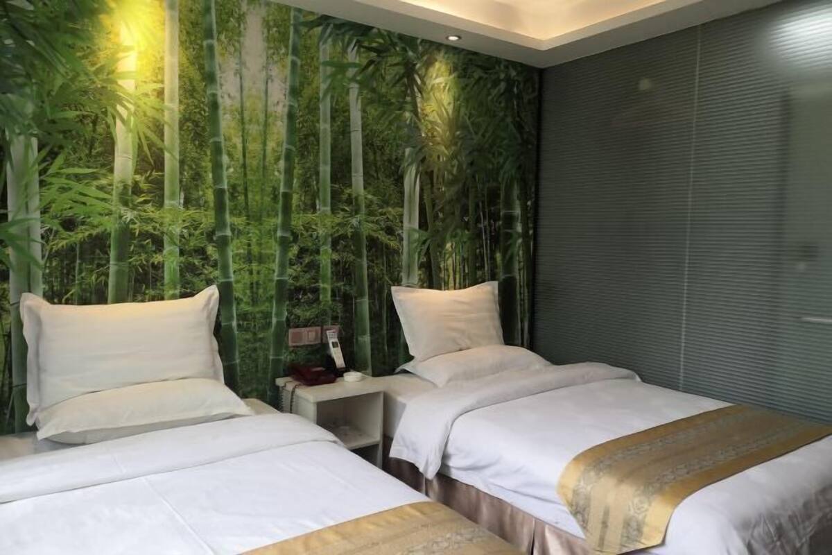 A hotel room with two beds, a wall mural of bamboo forest, a bedside table with a lamp, and a window with blinds.