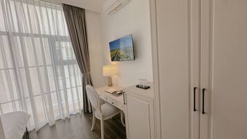 A hotel room with a desk, chair, and a bed. There is a window with curtains, an air conditioning unit, and a painting on the wall.