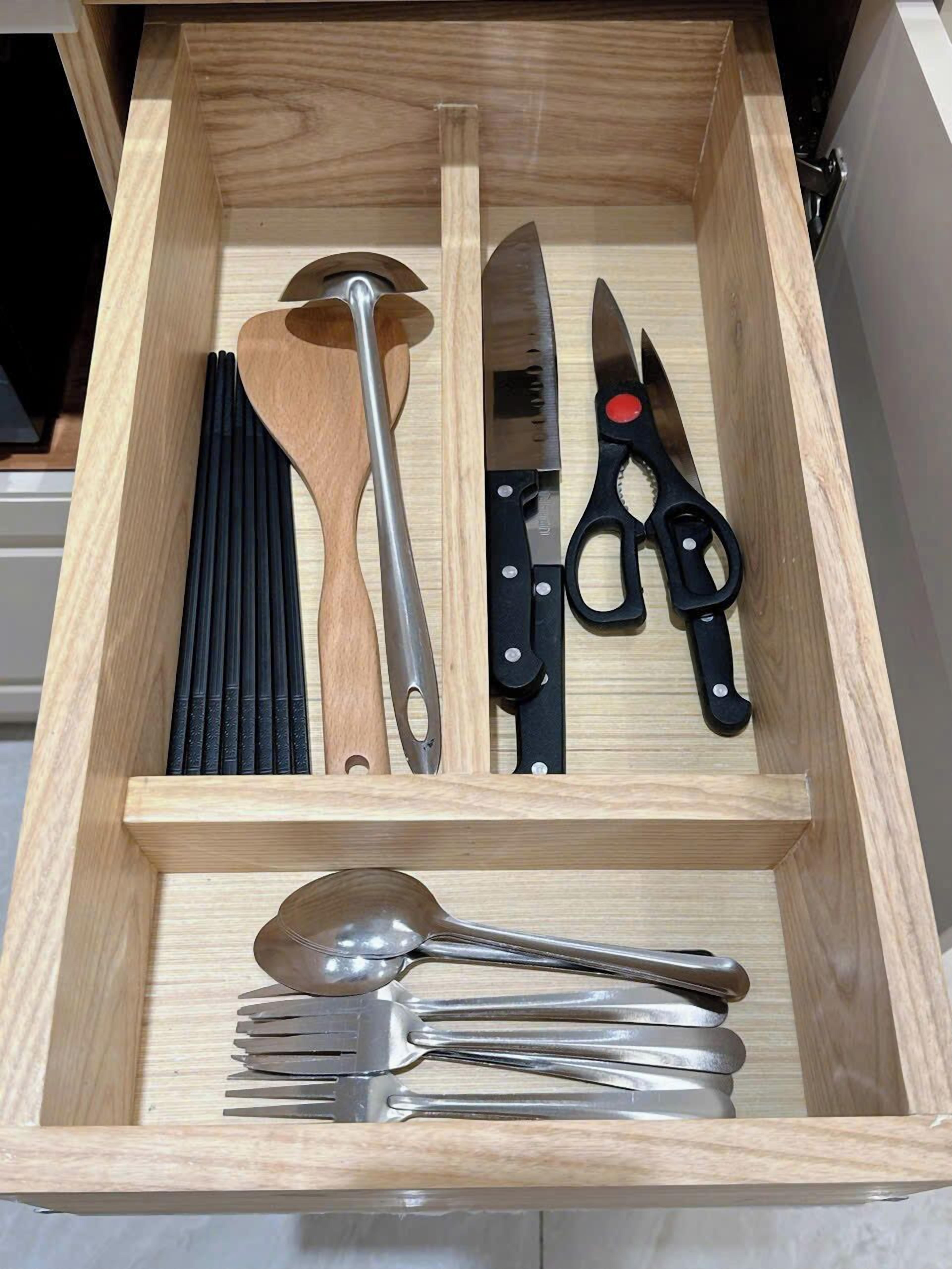 A drawer with various kitchen utensils including a wooden spoon, a large knife, a pair of scissors, and a set of forks.